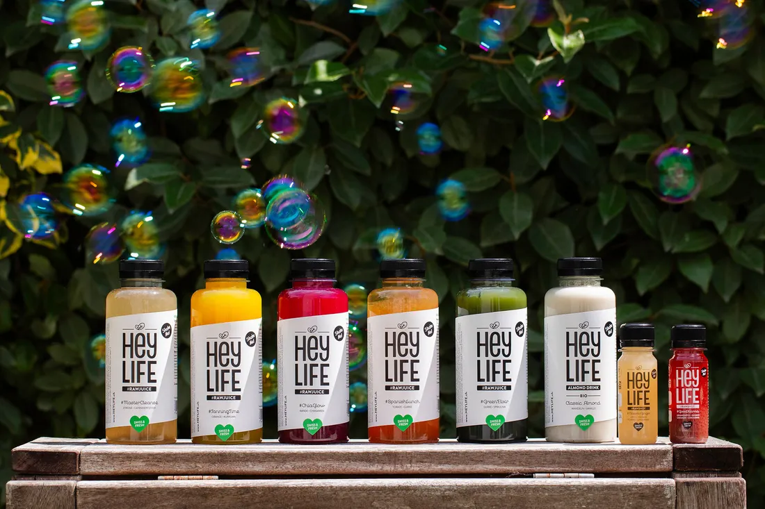 Heylife-Säfte sind in einer Linie aufgestellt, im Hintergrund ist eine Hecke mit grünen Blättern zu sehen. In der Luft schweben sanft Seifenblasen und schaffen eine verspielte Atmosphäre.