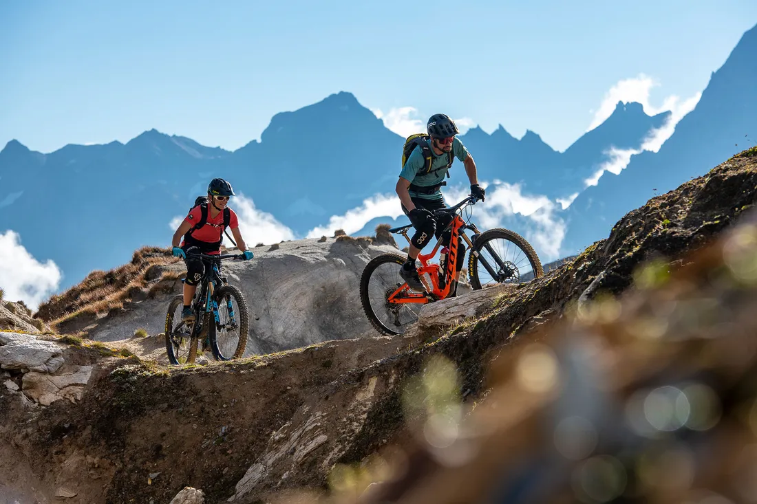 Zwei Mountainbiker fahren auf einem felsigen Bergpfad mit Berggipfeln im Hintergrund