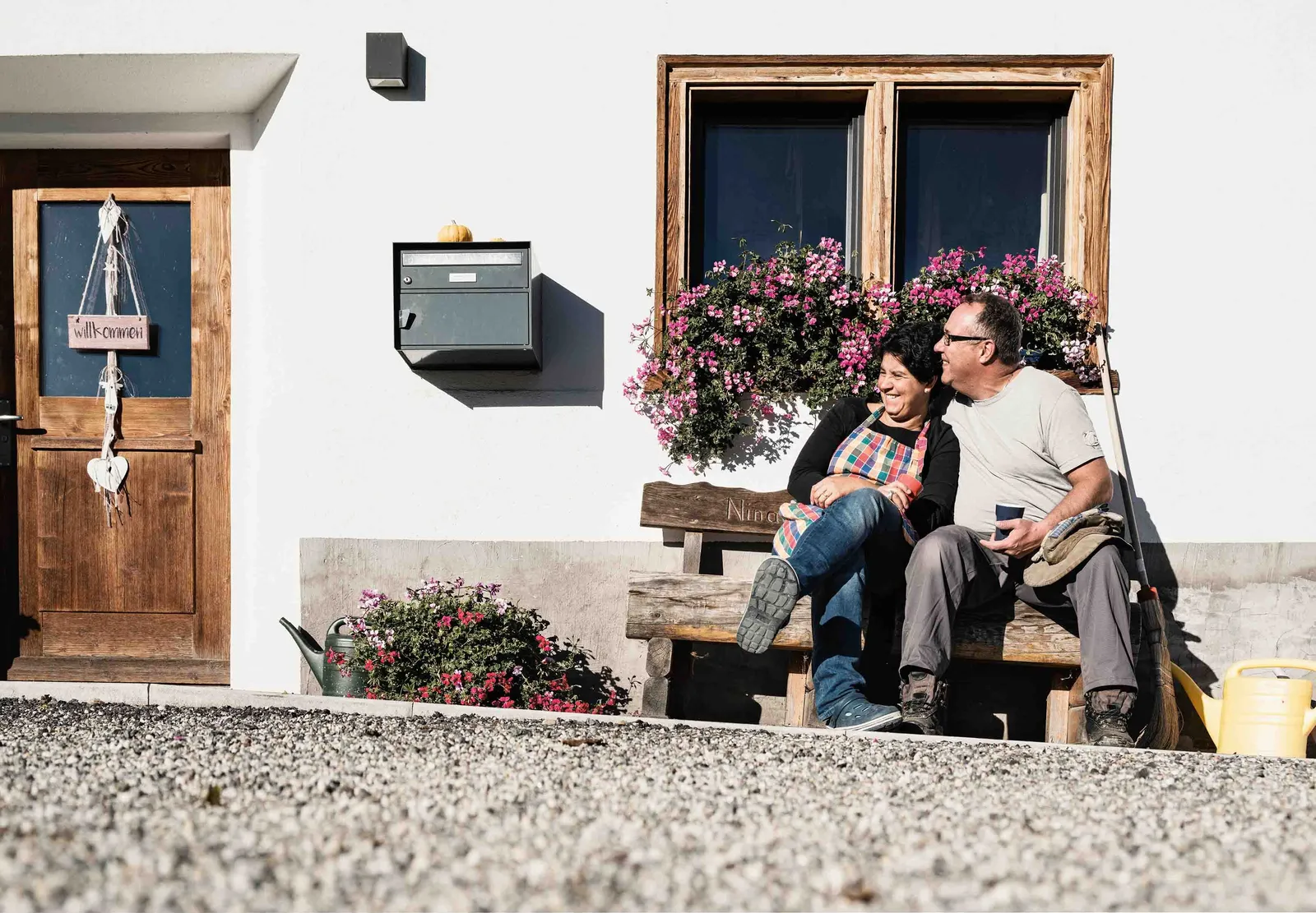 Due persone siedono su una panchina davanti a una casa con fiori e conversano rilassate.