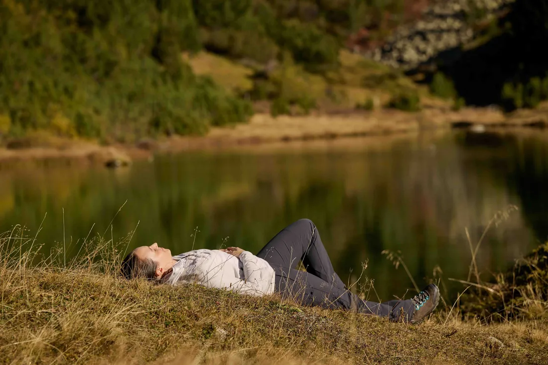 Eine Frau liegt im Gras neben einem See und tankt neue Energie.