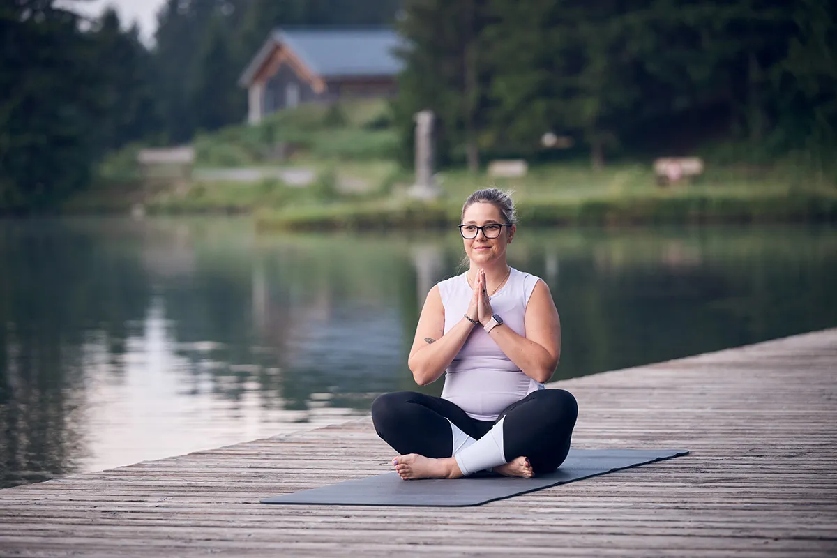 Eine junge Frau meditiert auf einem Holzsteg in der Nähe eines Sees.