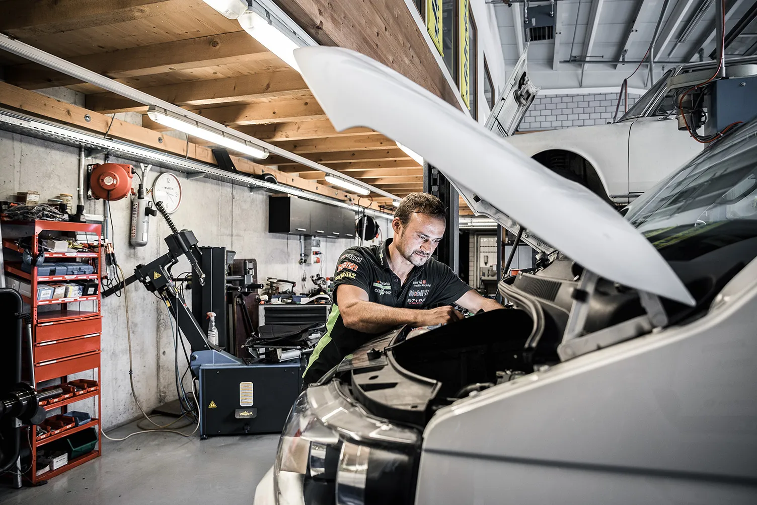Una persona lavora in un’officina automobilistica su un veicolo con il cofano aperto, circondata da attrezzi e apparecchi tecnici.