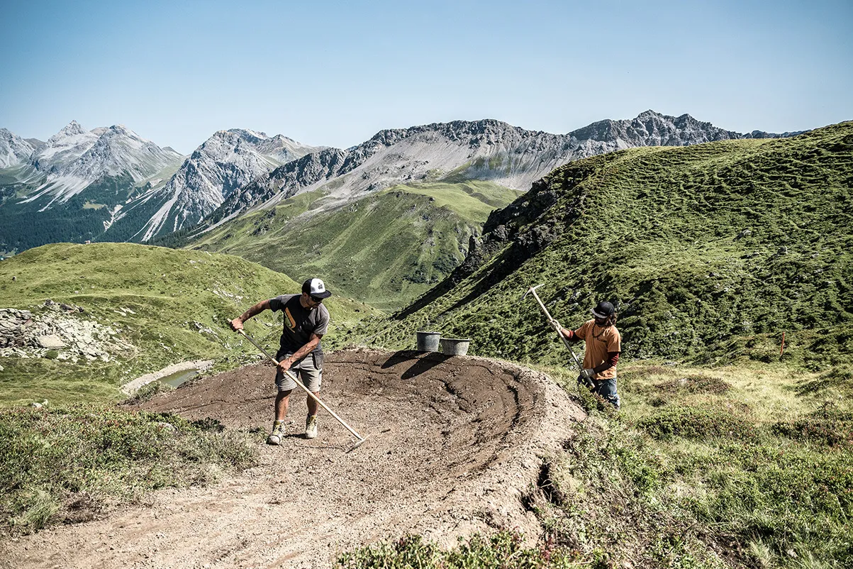 Due persone lavorano con attrezzi su un sentiero curvo in ambiente alpino, circondate da pendii verdi e montagne.