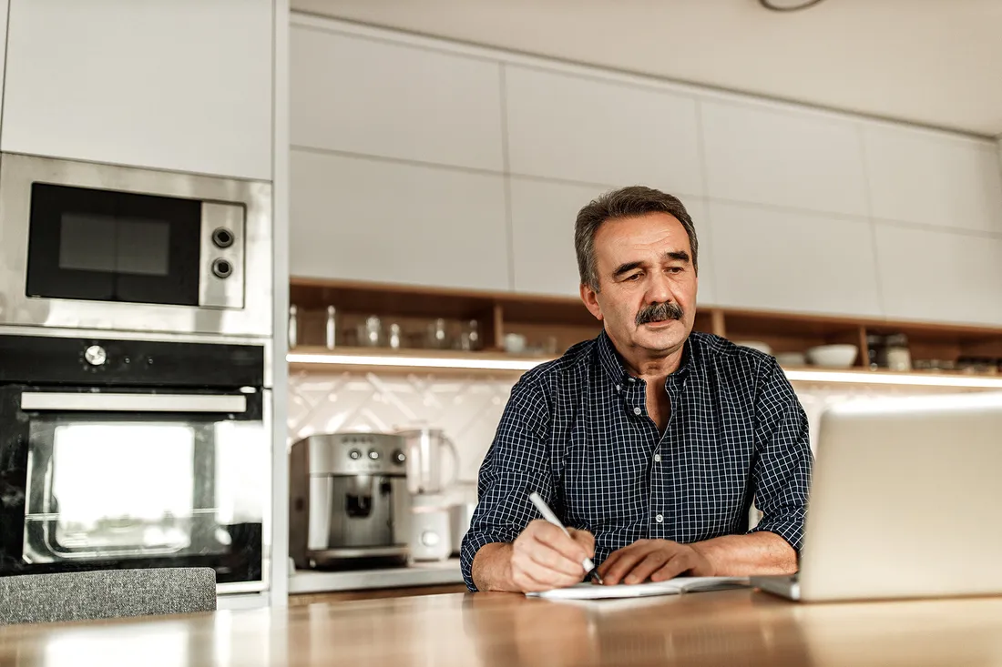 Una persona è seduta a un tavolo in cucina con un computer portatile e compila documenti scritti a mano.