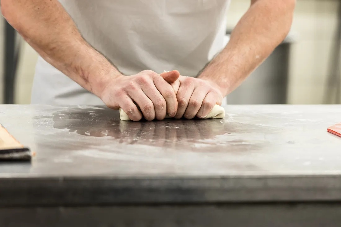 Delle mani impastano la pasta su una superficie di lavoro metallica in una cucina o panetteria professionale.