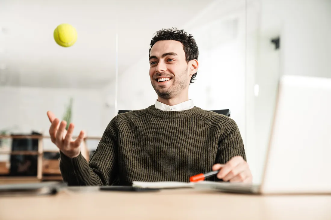 Una persona è seduta alla scrivania con un computer portatile e lancia una pallina da tennis come breve pausa di movimento in ufficio.