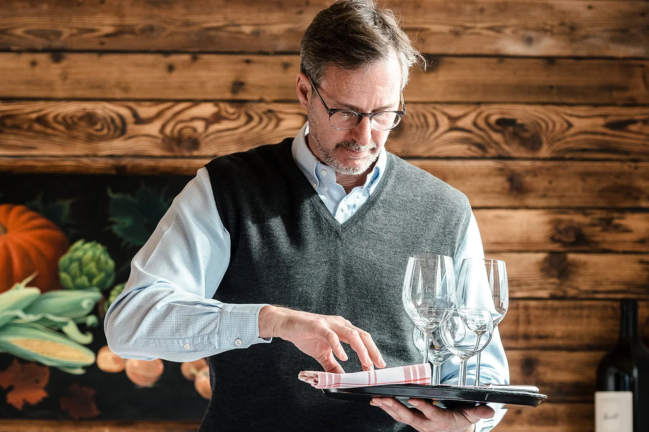 Une personne porte plusieurs verres à vin sur un plateau dans un restaurant, devant un mur en bois décoratif.