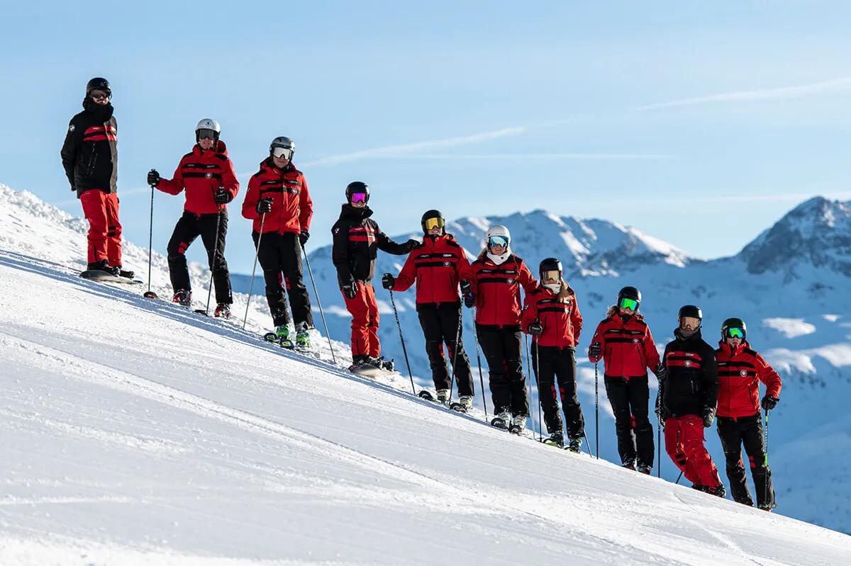 Zehn Skifahrer in roten Jacken stehen in einer Reihe auf einer schneebedeckten Piste mit Bergpanorama im Hintergrund