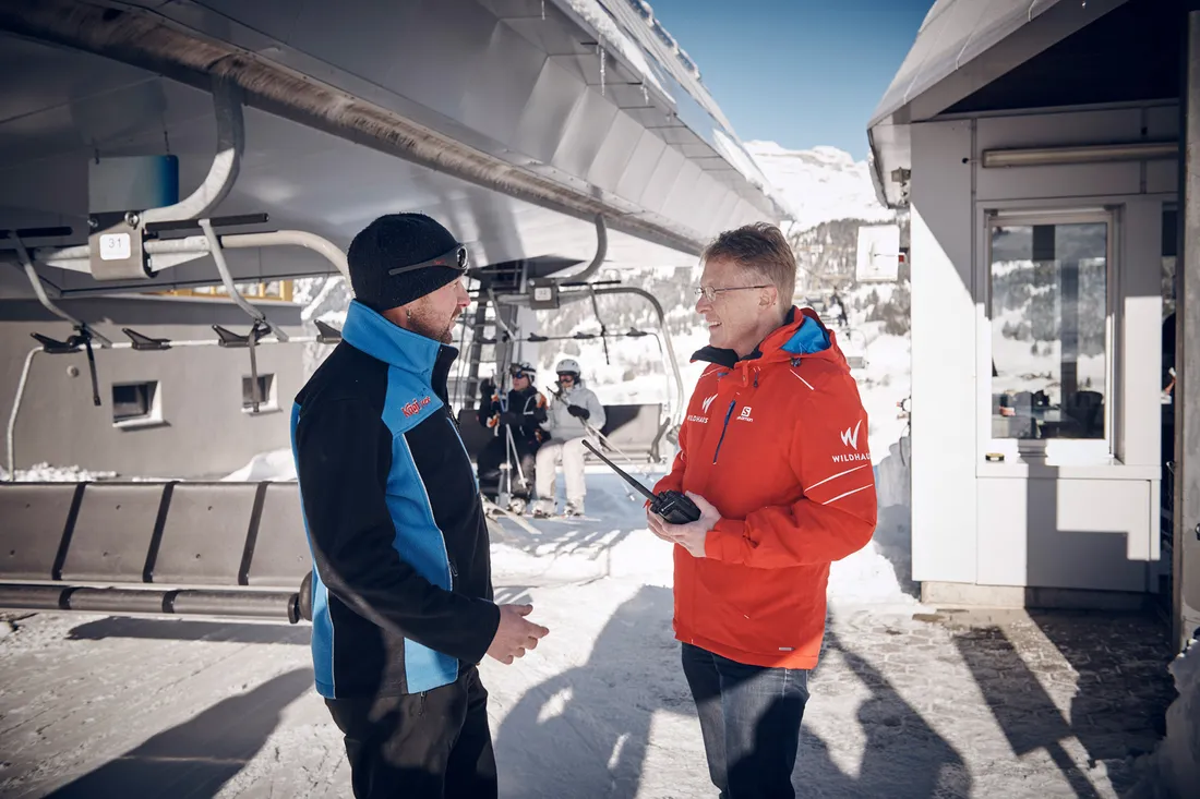 Zwei Männer in Winterjacken unter einer Skiliftstation im Schnee, einer hält ein Funkgerät