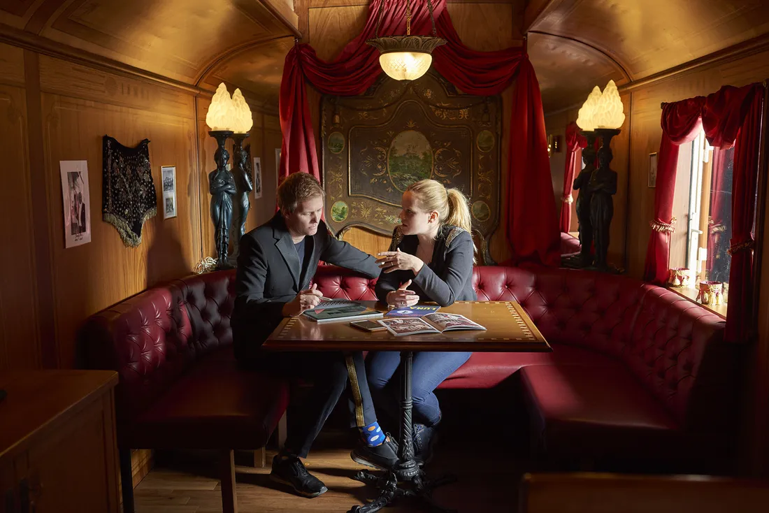 Zwei Personen sitzen an einem Holztisch in einem gemütlichen, mit rotem Samt dekorierten Raum und unterhalten sich