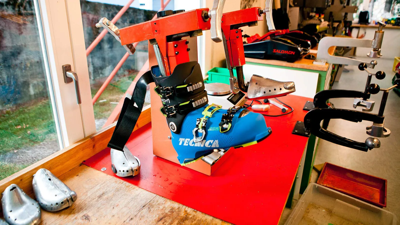 Blaue Tecnica Skischuhe in einer roten Skischuh-Bindungsvorrichtung in einer Werkstatt mit Werkzeugen und Schuhformen im Hintergrund