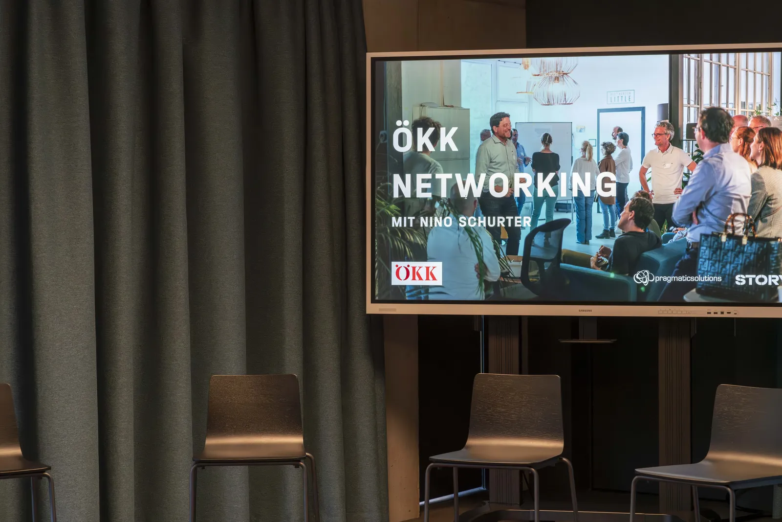 Großbildschirm zeigt 'ÖKK Networking mit Nino Schurter' vor Vorhang und leeren Stühlen in einem Veranstaltungsraum