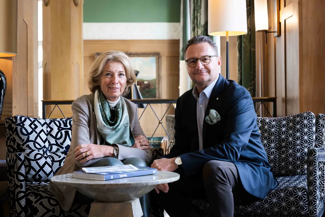 Zwei Personen sitzen in einem stilvollen Hotel-Lounge-Bereich auf Sesseln an einem kleinen Tisch mit Büchern und blicken zur Kamera.