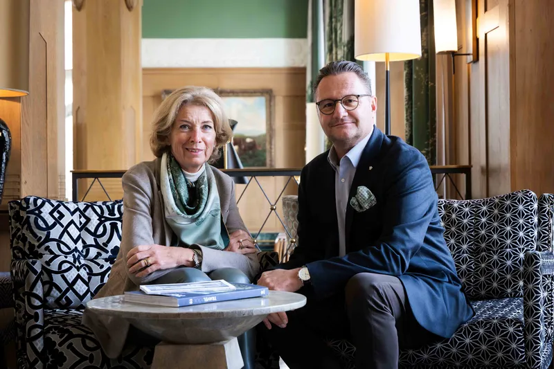 Zwei Personen sitzen in einem stilvollen Hotel-Lounge-Bereich auf Sesseln an einem kleinen Tisch mit Büchern und blicken zur Kamera.