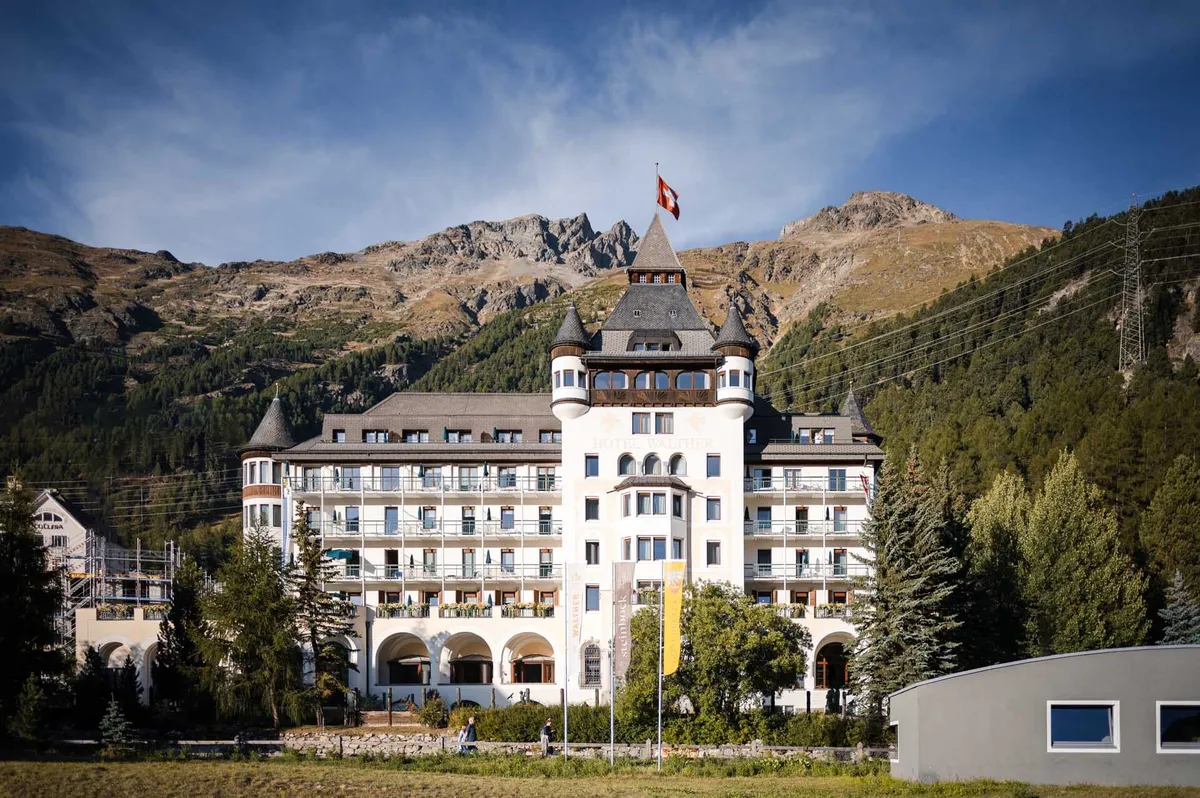 Ein historisches Hotelgebäude mit Türmen steht vor einer alpinen Bergkulisse mit Schweizer Flagge auf dem Dach.