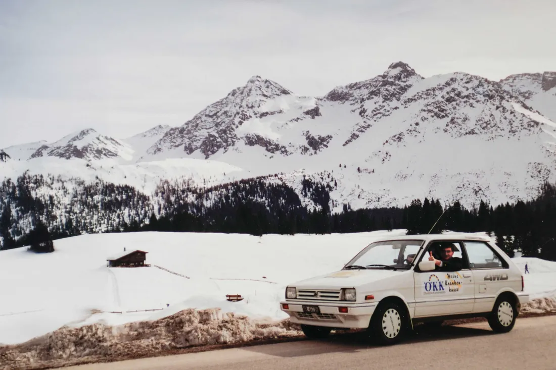 Weisser ÖKK-Dienstwagen mit altem „Meine Krankenkasse“-Logo vor verschneiter Alpenlandschaft; der Fahrer winkt aus dem Fenster.