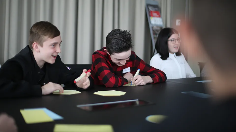 Drei Jugendliche sitzen an einem Tisch, zwei lachen und einer schreibt etwas auf Papier.