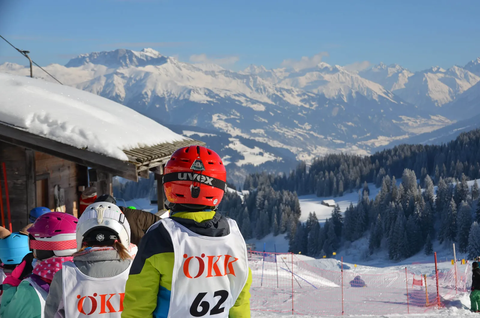 Kinder in bunter Skiausrüstung mit Helmen und Nummern auf Rücken stehen vor verschneitem Bergpanorama