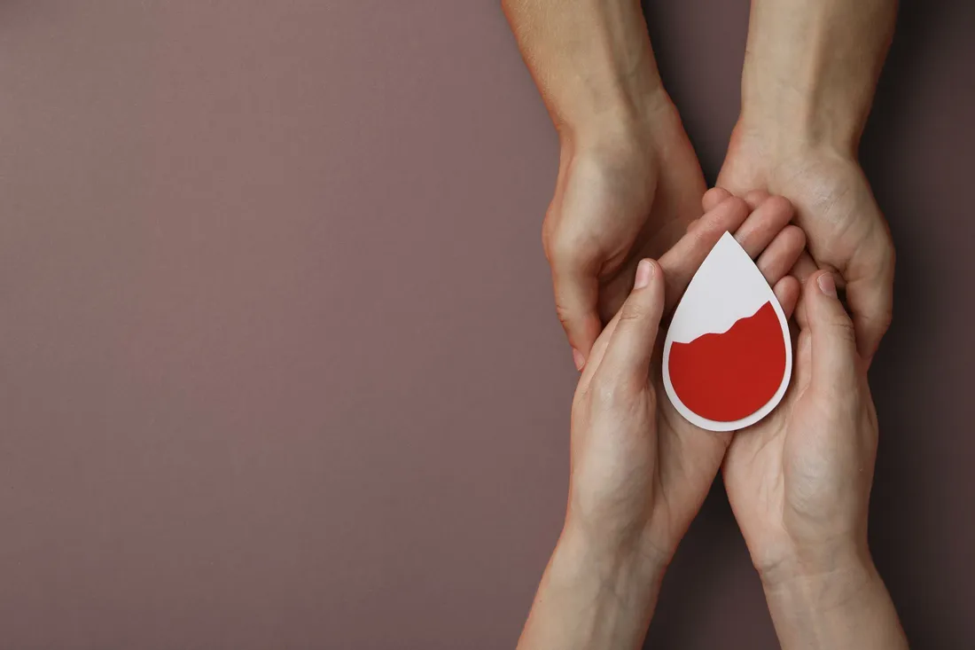 Zwei Hände halten eine weitere Hand, die eine Papierform in Tropfenform mit rotem Inhalt hält.