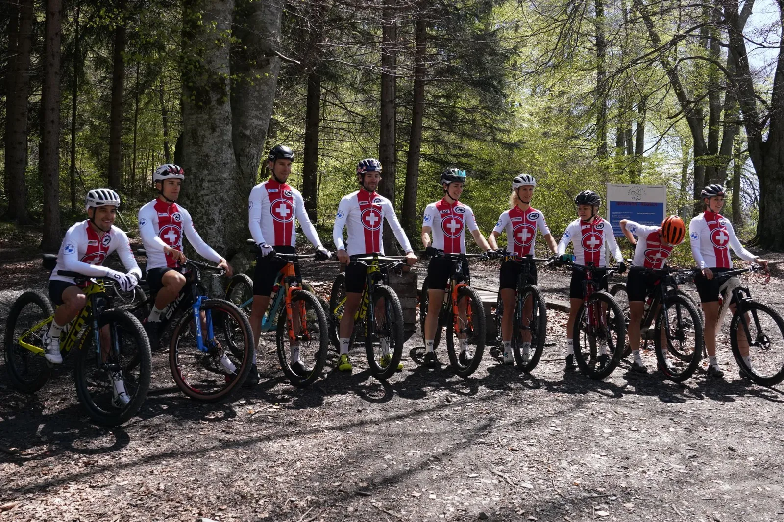 Neun Radfahrer in weiß-roten Trikots stehen mit ihren Mountainbikes auf einem Waldweg im Sonnenschein