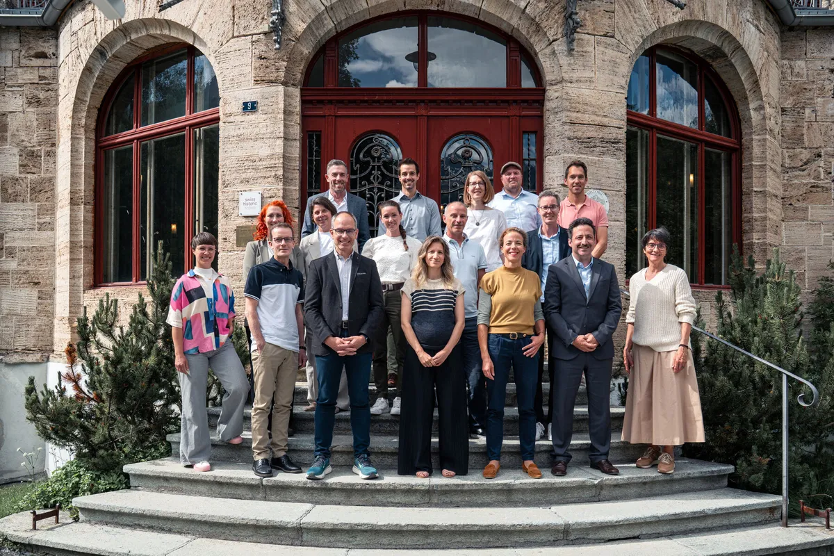 Il consiglio di fondazione della Fondazione ÖKK è riunito in un gruppo numeroso su una scalinata davanti a un edificio storico con finestre ad arco.
