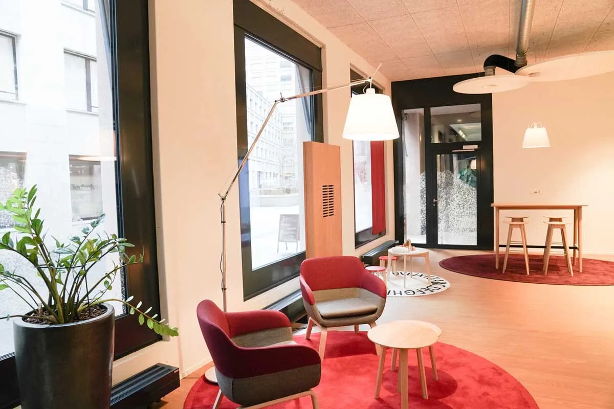 Modernes Agenturbüro mit roten Sesseln, Stehlampe, kleinen Tischen und grossen Fenstern, helle und offene Gestaltung.