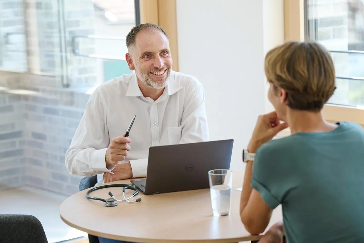 Ein Arzt im Gespräch mit einer Patientin in einem hellen, modernen Sprechzimmer. Auf dem Tisch liegen ein Laptop, ein Stethoskop und ein Glas Wasser.