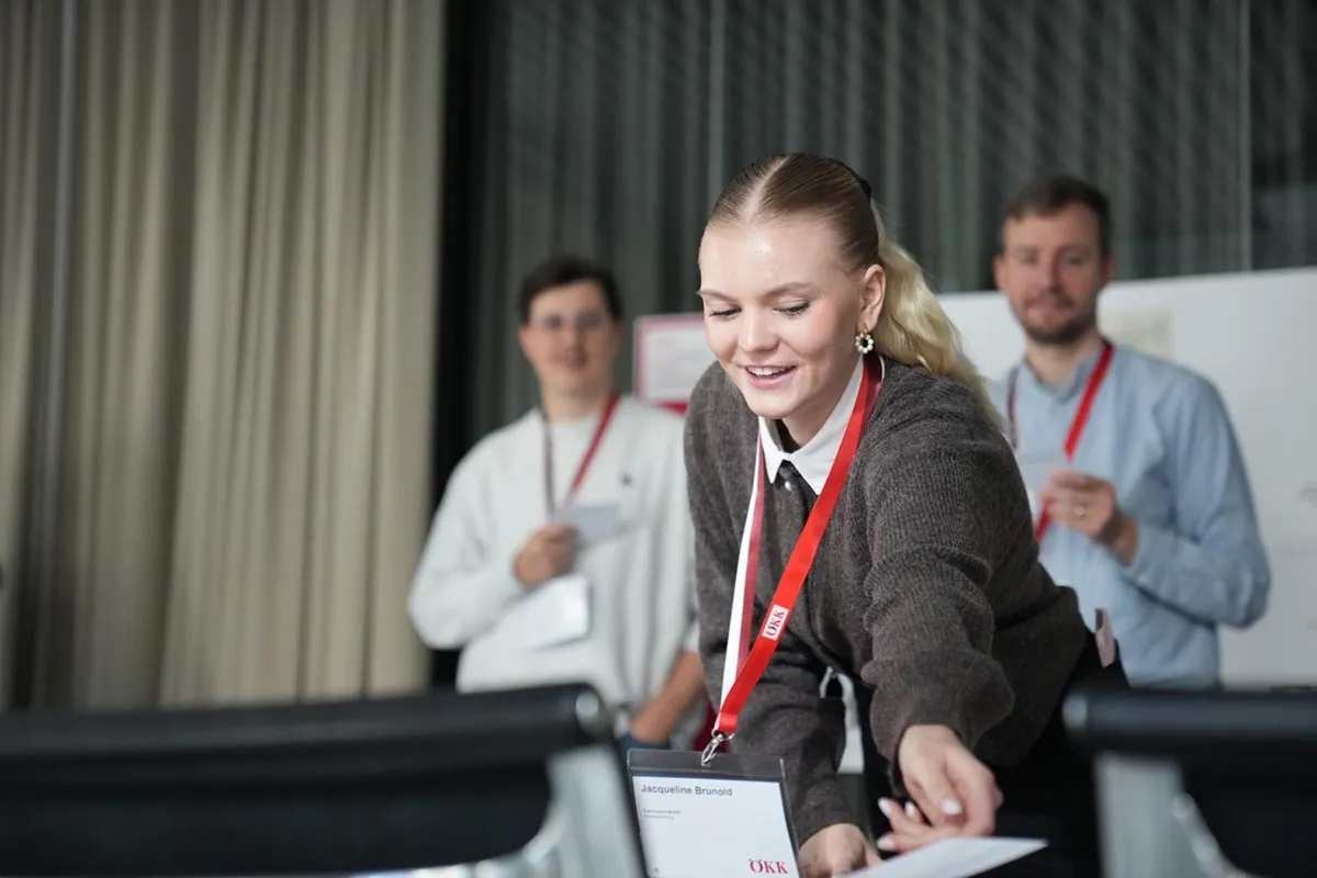 Mitarbeitende beim ÖKK-Welcomeday: Eine junge Frau mit Namensschild und rotem Lanyard verteilt Unterlagen, im Hintergrund stehen zwei weitere Kollegen.