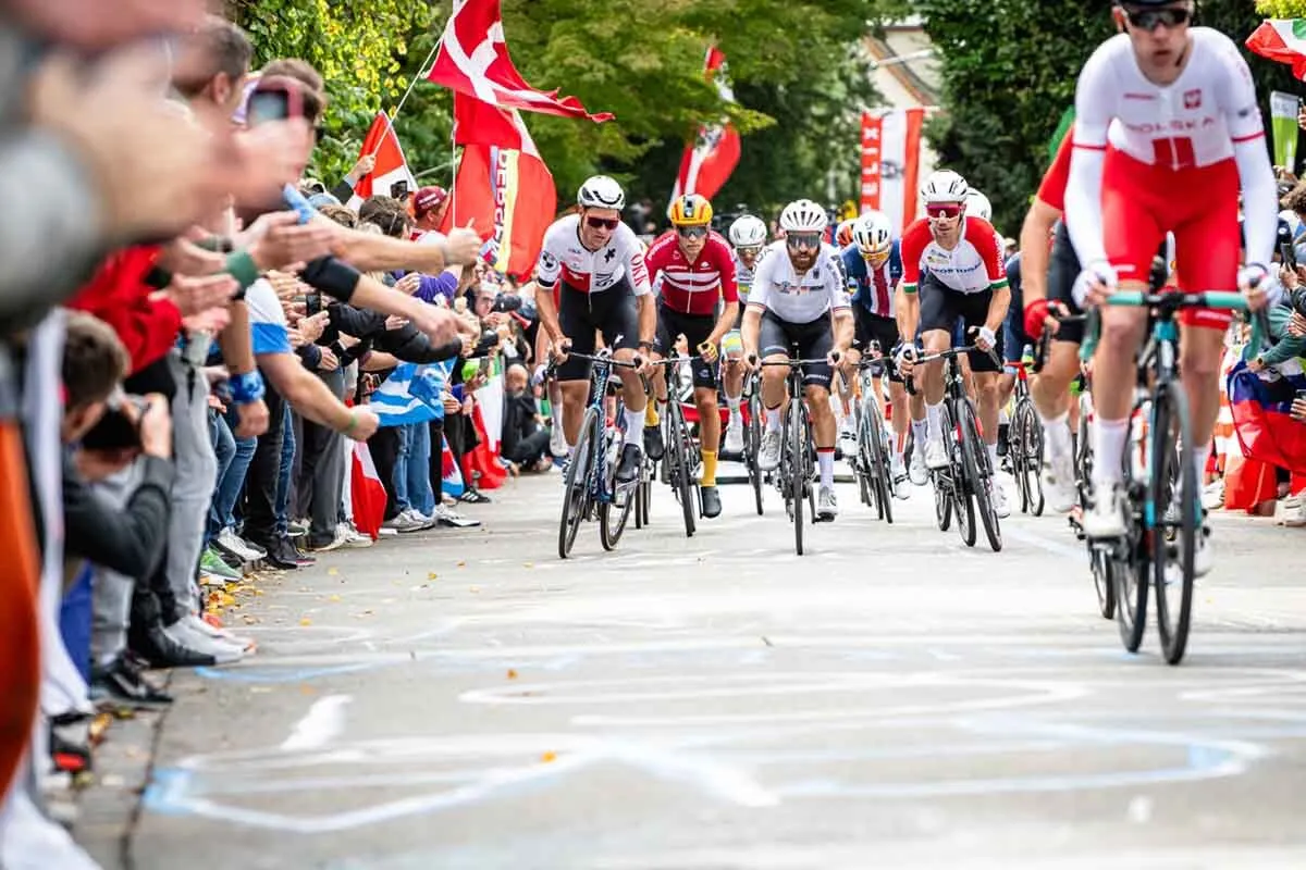 Radrennfahrer bei einem internationalen Wettbewerb fahren durch eine enge Straße, gesäumt von jubelnden Zuschauern mit Fahnen und Kameras. Im Vordergrund sind Fahrer in Nationaltrikots zu sehen, darunter Deutschland, Dänemark und Polen. Die Stimmung ist dynamisch und energiegeladen.