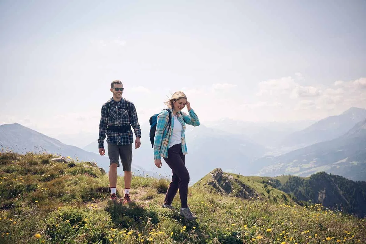 Zwei Wanderer, ein Mann und eine Frau, gehen auf einem grasbewachsenen Berggipfel mit Blick auf ein Tal und Berge im Hintergrund, sonniger Tag.
