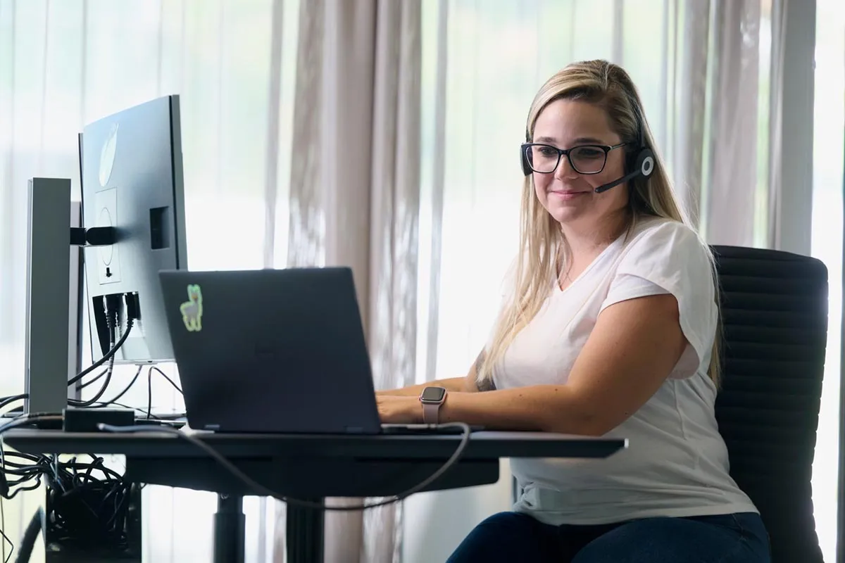 Frau mit Headset und Brille arbeitet an Laptop und großem Monitor in modernem Büro von ÖKK.