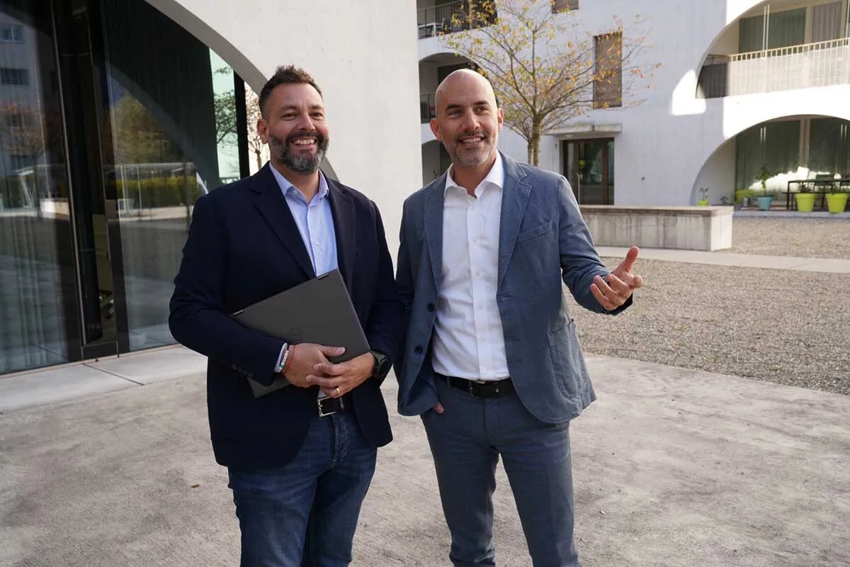 Zwei Männer in Anzügen stehen vor einem modernen Gebäude - dem Hauptsitz von ÖKK, einer hält einen Laptop, sie unterhalten sich freundlich.