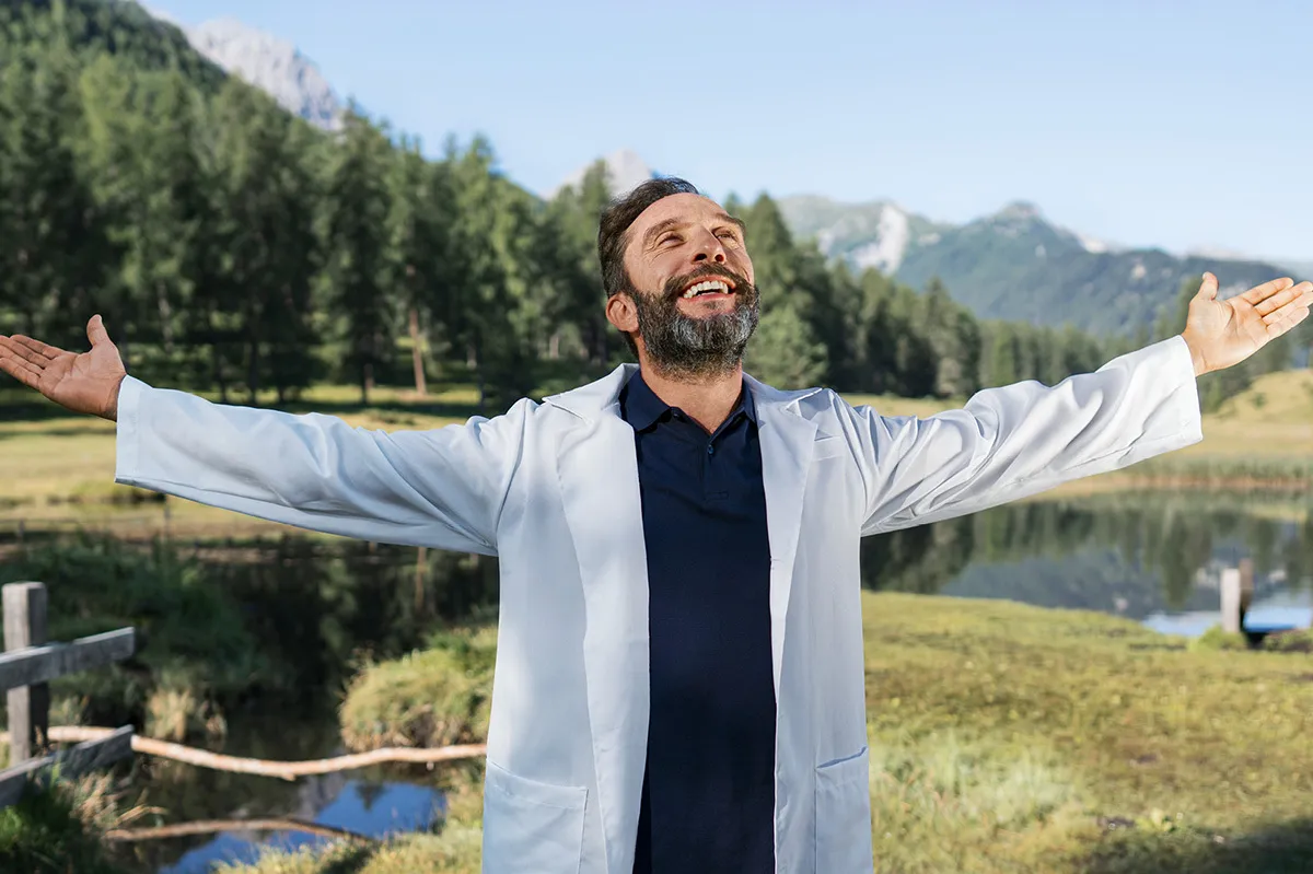 Ein Mann in einem weissen Kittel steht in einer bergigen Landschaft, mit ausgestreckten Armen und einem glücklichen Gesichtsausdruck und geniesst offensichtlich die frische Bergluft.