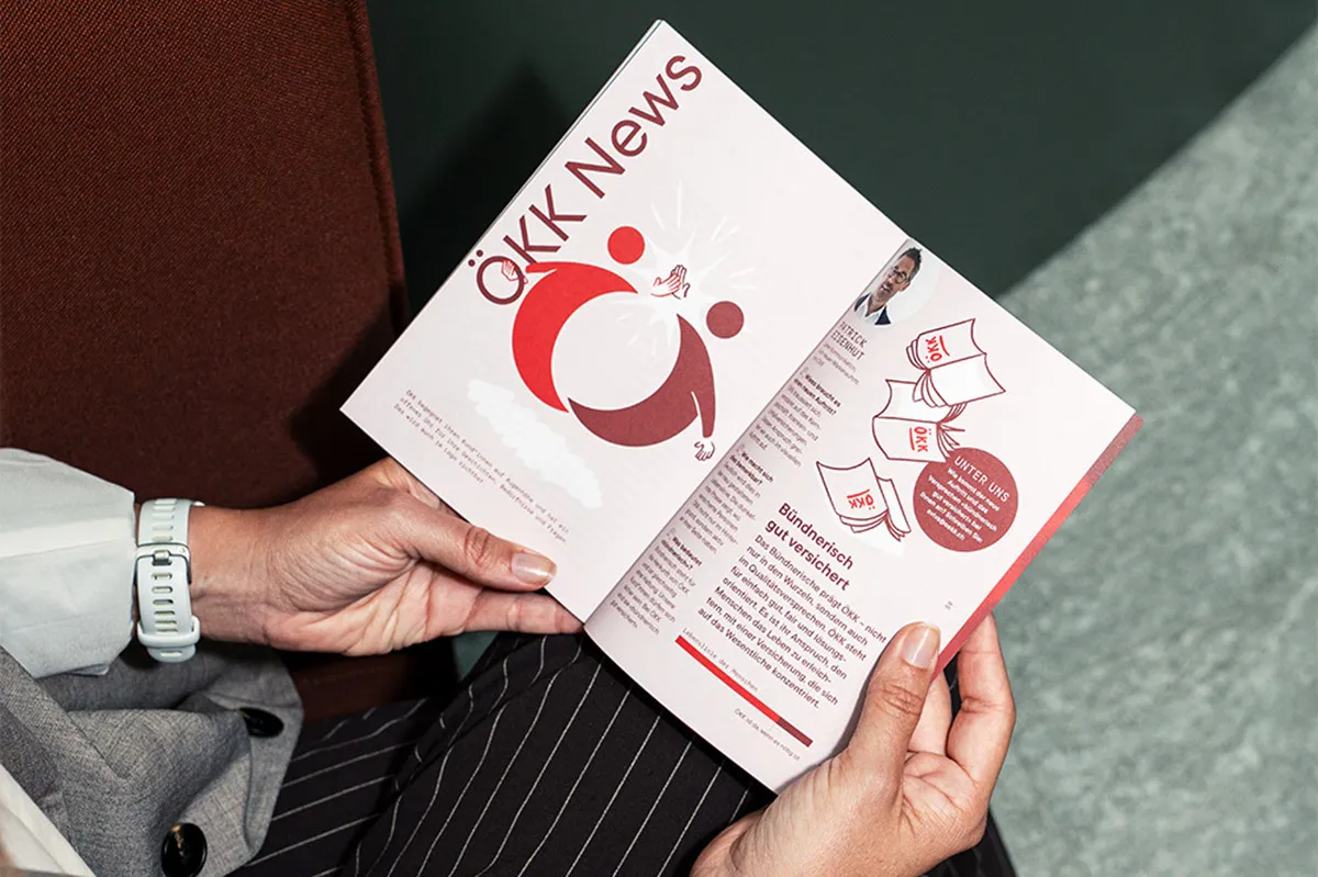Una persona tiene tra le mani la rivista clienti ÖKK News aperta, con articoli e illustrazioni nel design ÖKK.