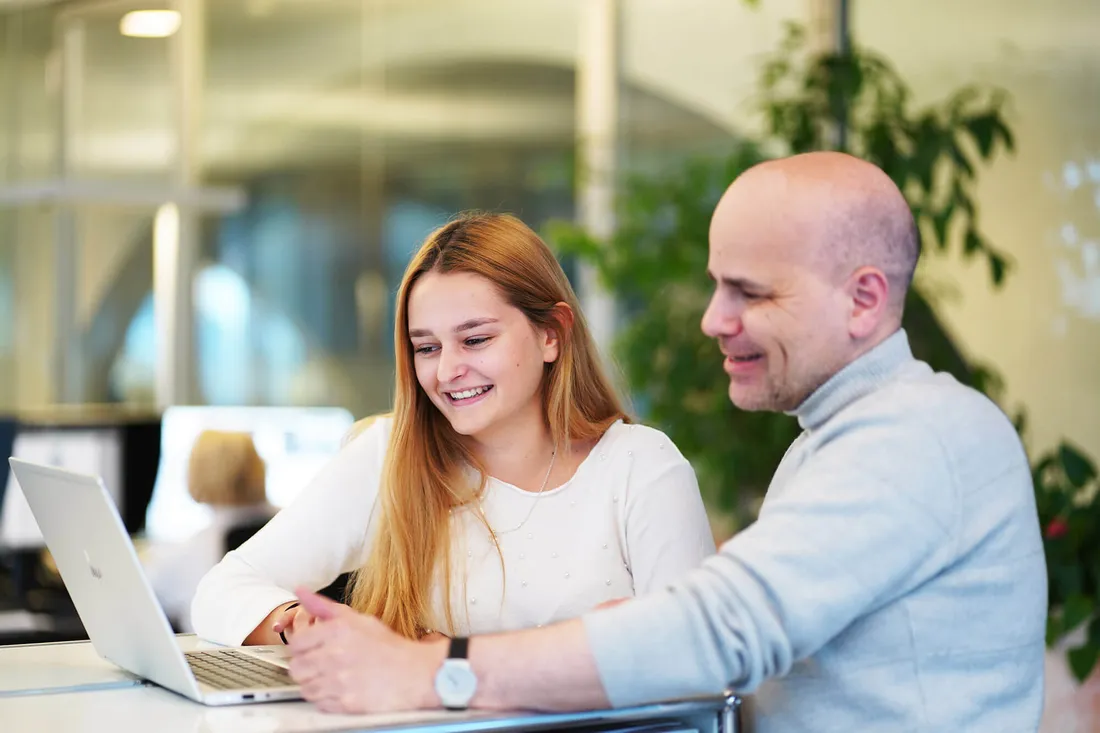 Zwei Personen, eine junge Frau und ein Mann, arbeiten gemeinsam an einem Laptop in einem modernen Büro