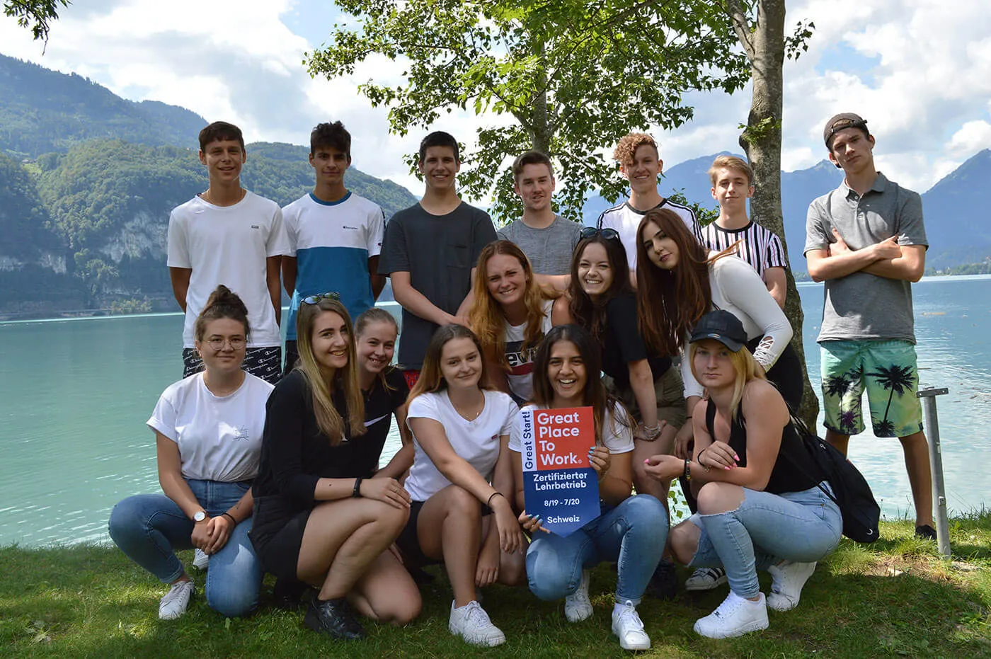 Gruppo di 14 giovani che sorridono e si mettono in posa sulla riva di un lago davanti a montagne e alberi
