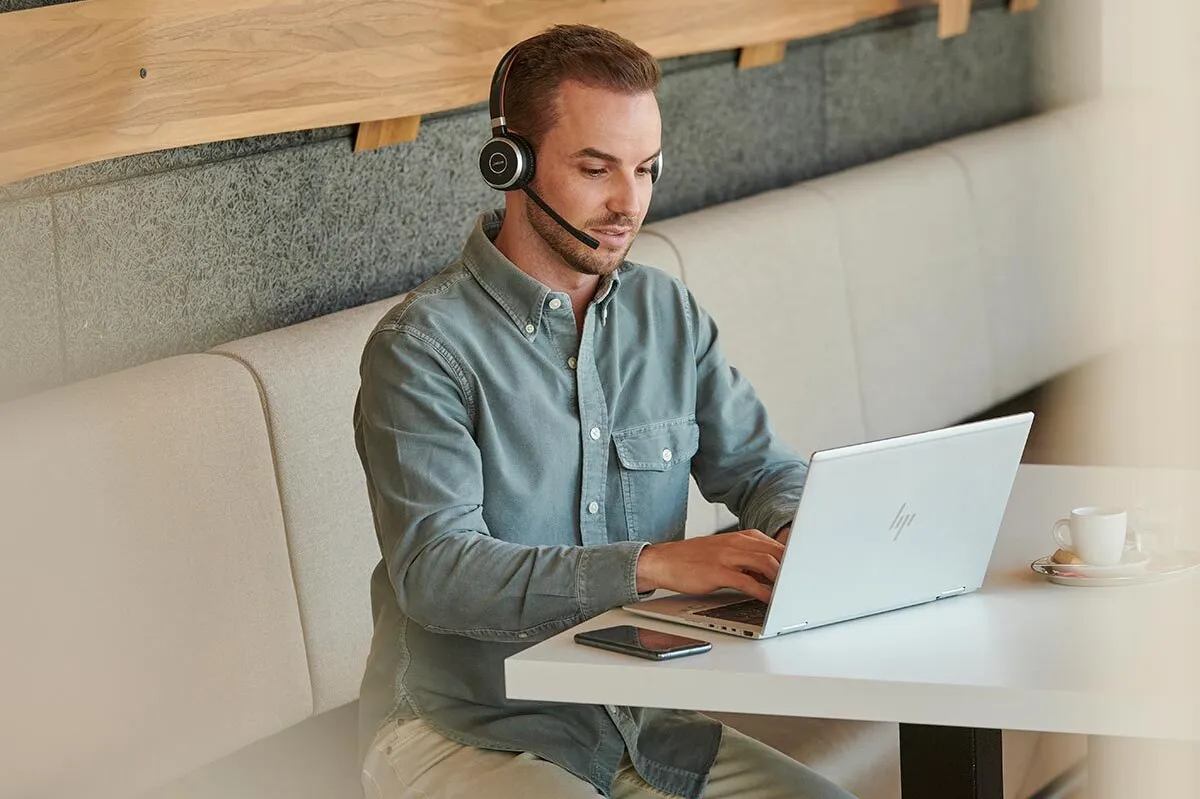 Mann mit Headset arbeitet an einem Laptop in einem Café, neben ihm liegen Smartphone und Kaffeetasse