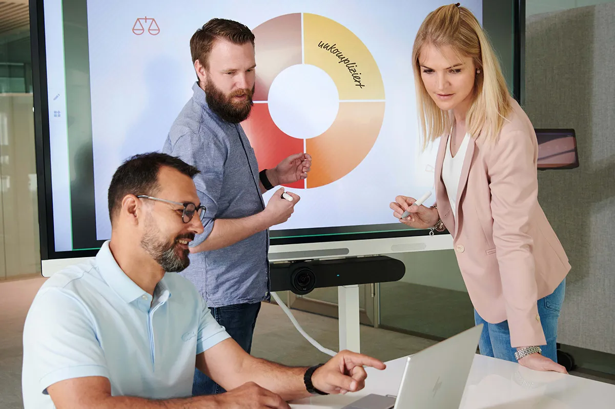 Drei Personen diskutieren vor einem Bildschirm mit einem farbigen Kreisdiagramm, eine Frau zeigt auf das Diagramm, zwei Männer hören zu.