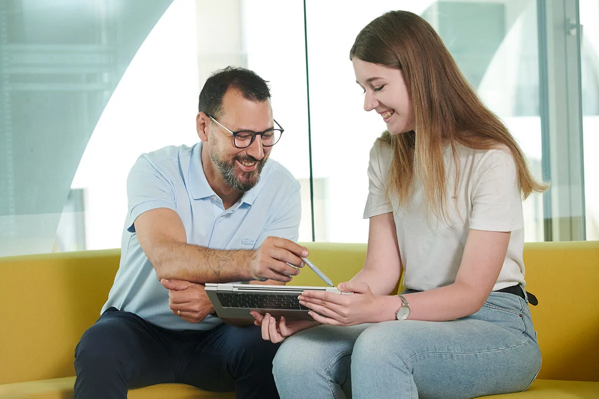 Zwei Personen sitzen auf einem gelben Sofa, schauen lächelnd auf ein Tablet und diskutieren etwas
