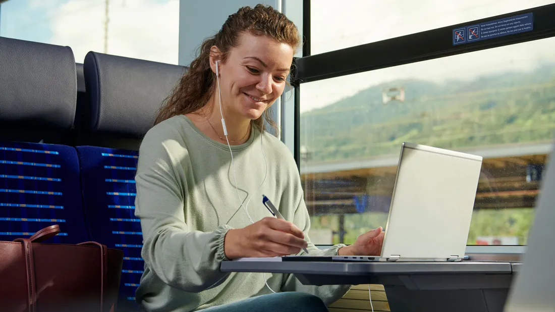 Junge Frau mit Ohrhörern arbeitet lächelnd an Laptop und macht Notizen im Zug mit Landschaft im Hintergrund