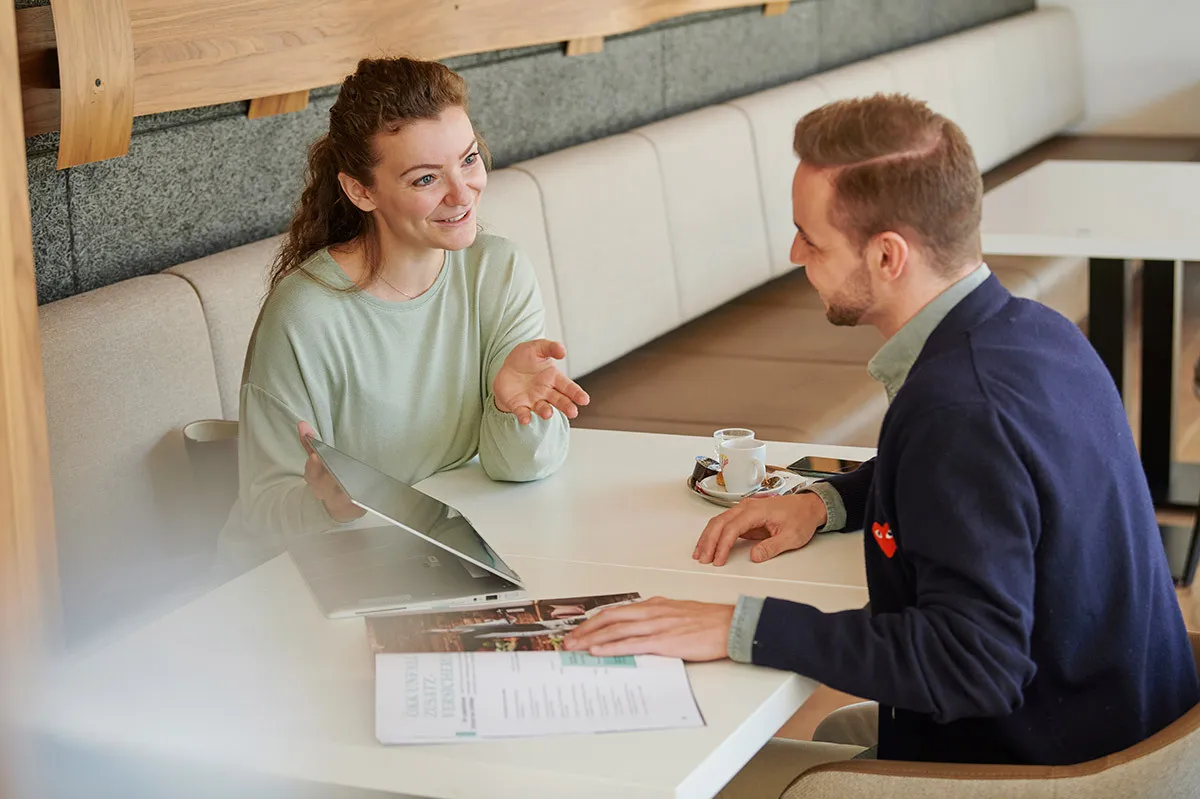 Zwei Personen sitzen an einem Tisch und führen ein Gespräch, eine Frau erklärt etwas mit Gestik, ein Mann hört aufmerksam zu