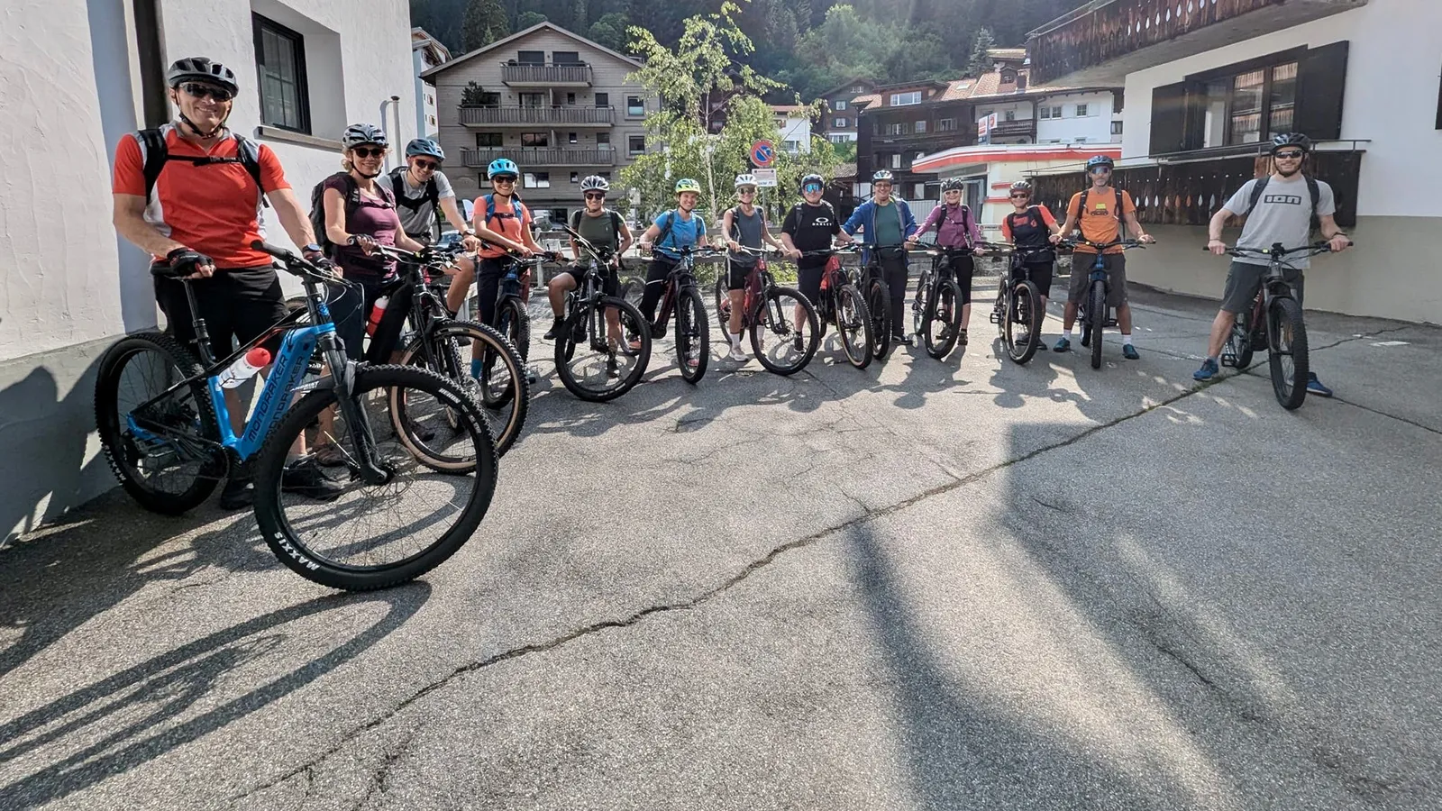 Gruppe von dreizehn Radfahrern mit Helmen und Sportkleidung steht mit ihren Fahrrädern auf einer Straße in einem Dorf mit Bergen im Hintergrund