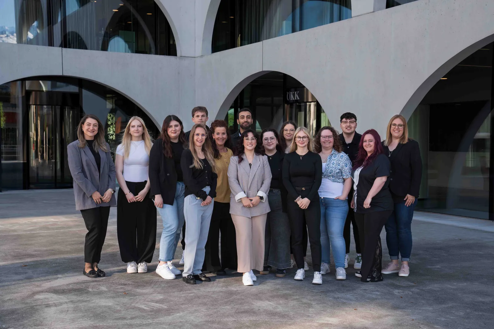 Gruppenfoto eines vielfältigen Teams vor einem modernen Bürogebäude mit dem Schriftzug ÖKK. Die Gruppe steht für moderne Führung und Leadership im Sinne des PERMA-Lead-Modells – mit Fokus auf positive Psychologie, Teamdynamik und stärkenorientierter Zusammenarbeit.