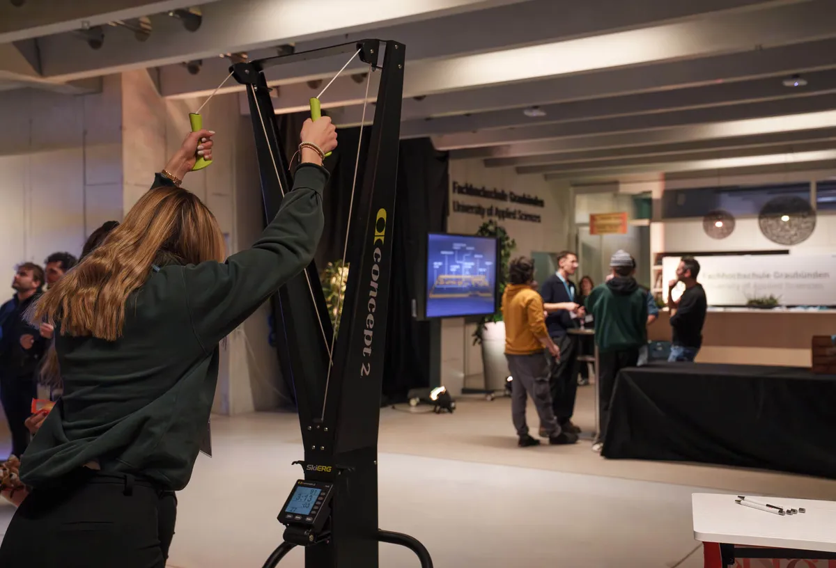 Person mit langem Haar zieht an Griffen eines schwarzen Fitnessgeräts der Marke Concept2 in einem Innenraum mit mehreren Personen im Hintergrund.
