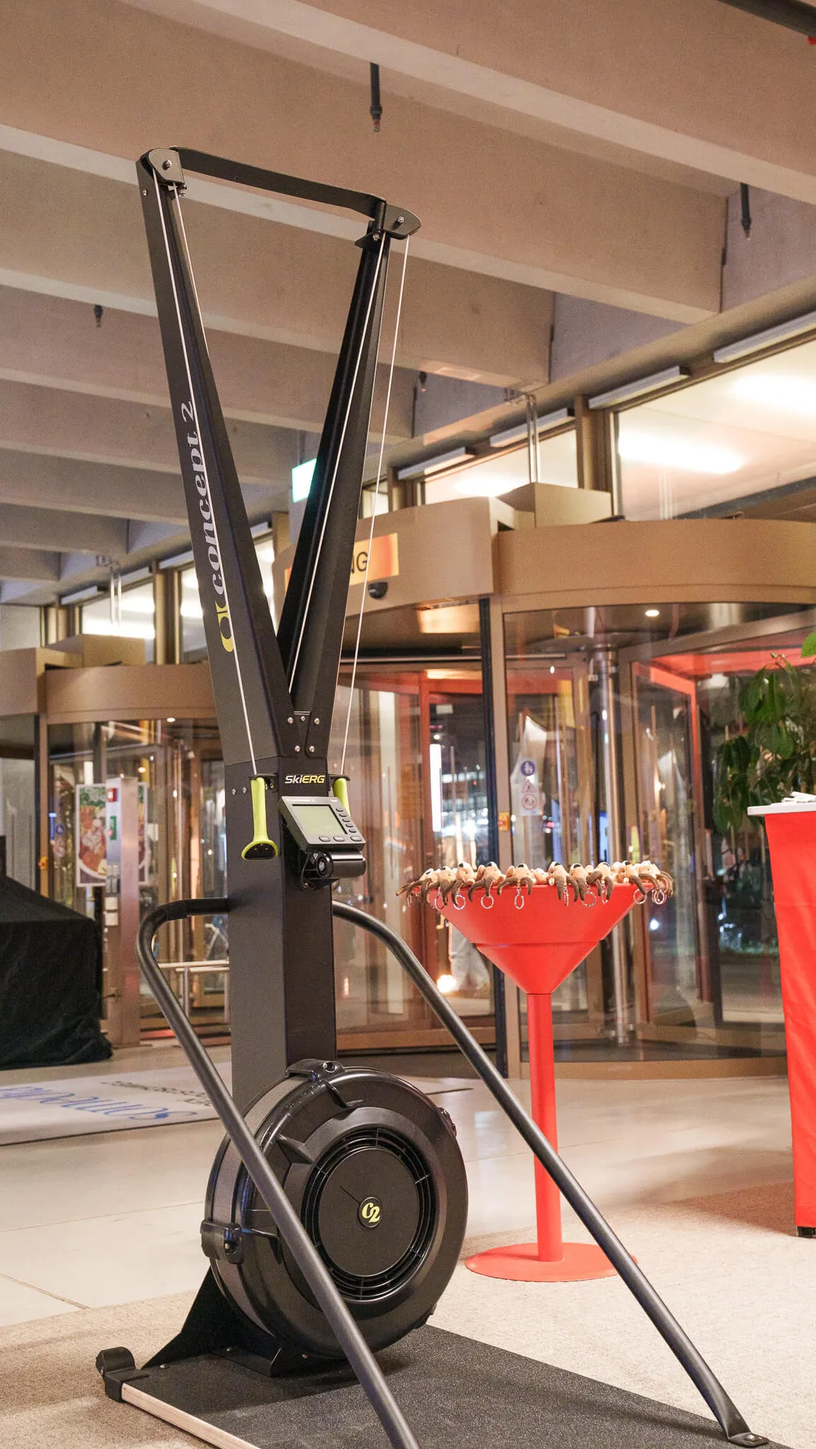 Rudergerät mit schwarzem Rahmen und gelben Akzenten steht in einem Innenraum mit Glasdrehtür und roten Stehtischen.