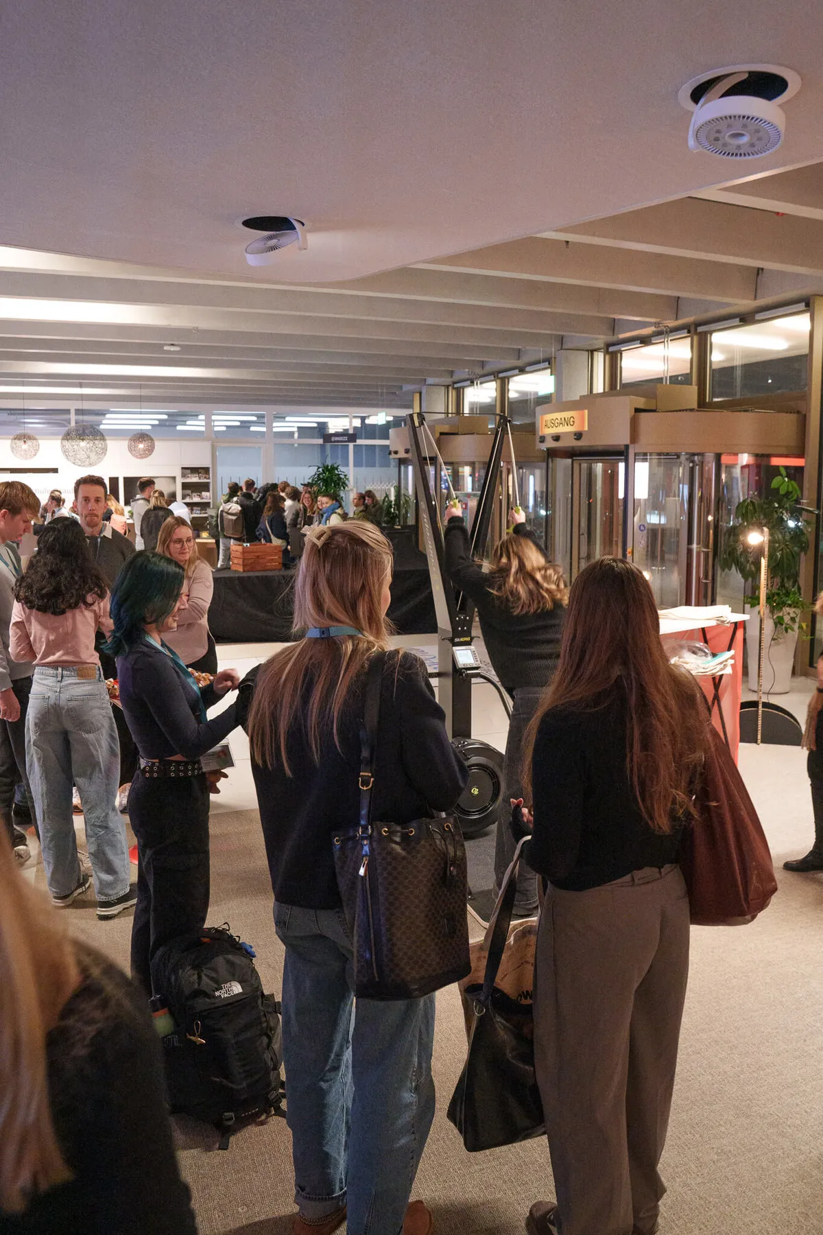 Mehrere Personen stehen in einem hell beleuchteten Innenraum mit sichtbaren Deckenbalken und großen Fenstern, einige tragen Rucksäcke und Taschen.