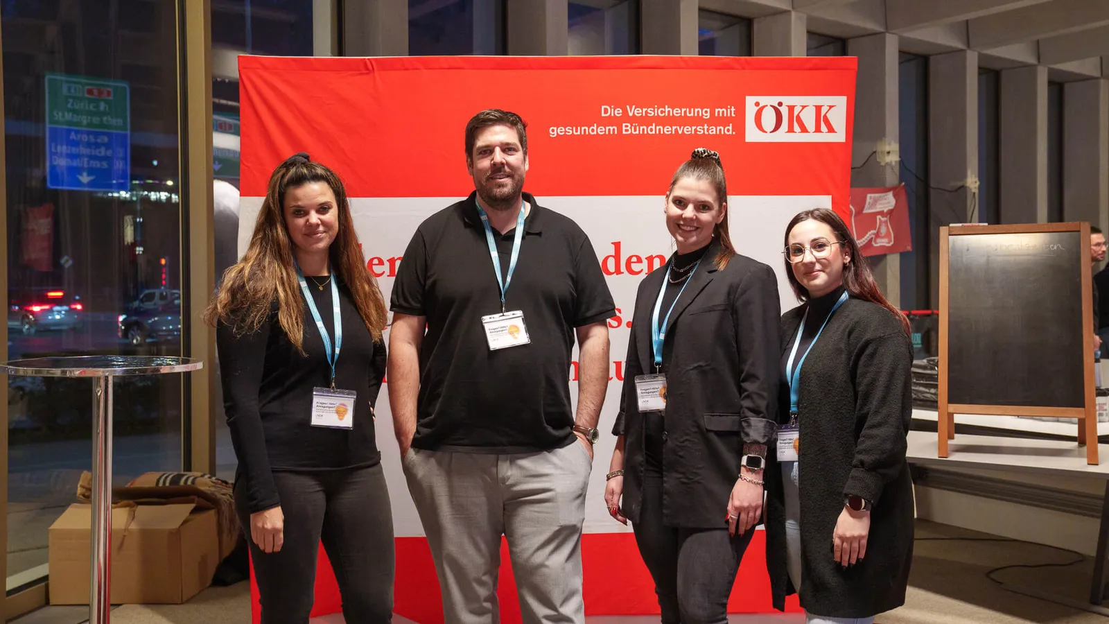 Vier Personen mit Namensschildern stehen vor einem roten und weißen ÖKK-Werbeplakat in einem Innenraum.