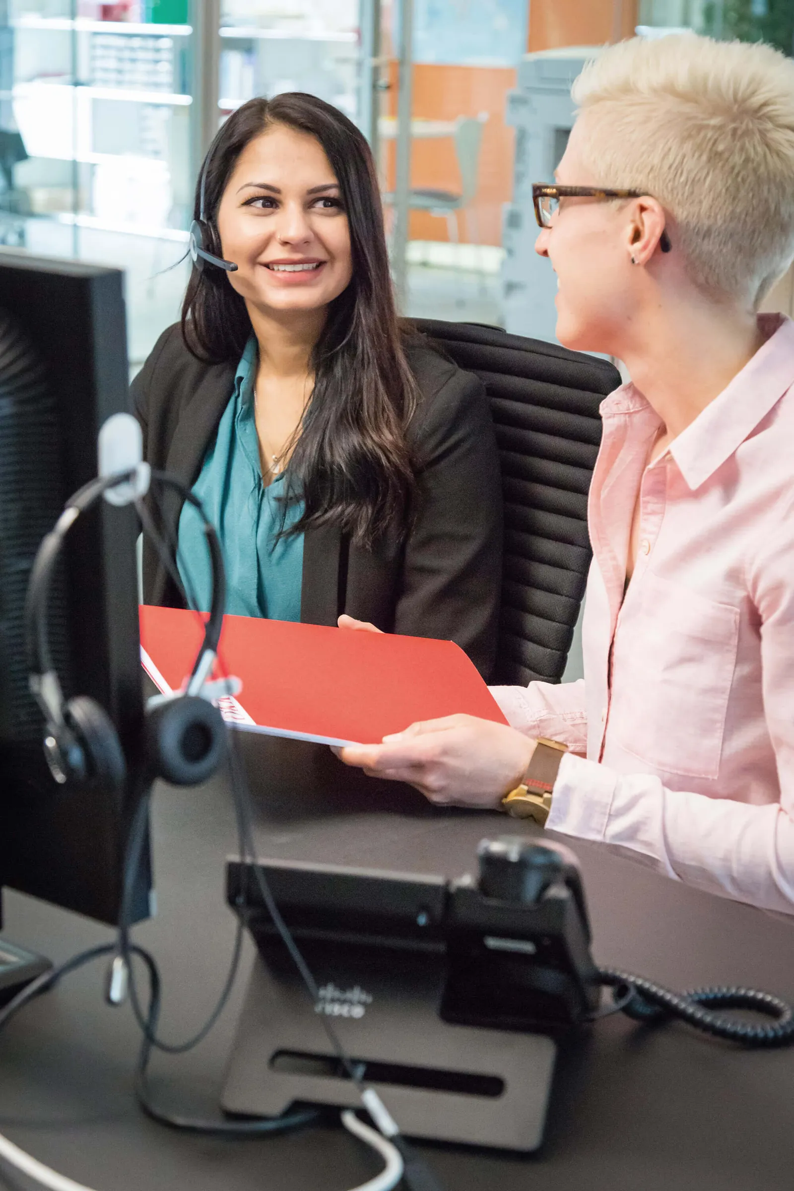 Zwei Frauen im Gespräch an einem Schreibtisch mit Headset und Dokumenten, eine hält einen roten Ordner