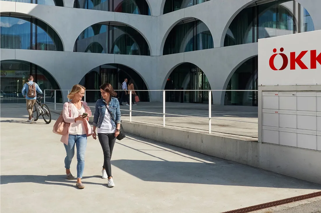 Zwei Frauen gehen vor dem ÖKK Hauptsitz mit bogenförmiger Fassade und schauen gemeinsam auf ein Smartphone.